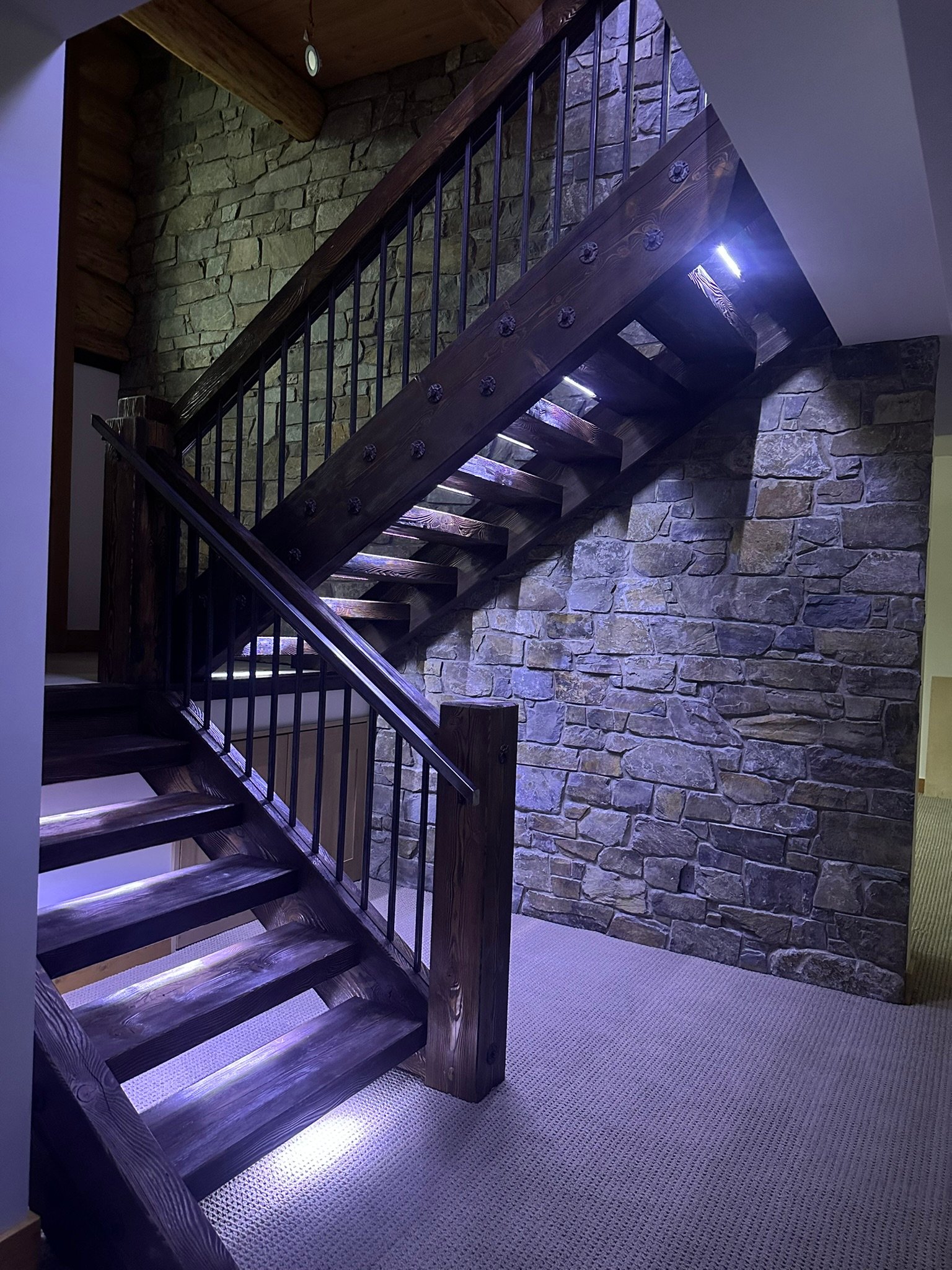 Interior photograph of a luxury Kootenay home addition featuring a custom heavy timber staircase with integrated LED lighting metal spindles and a beautiful natural stone accent wall.
