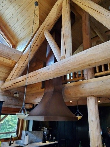 Interior photograph of a luxury Kootenay log home kitchen featuring massive natural round wood beams framing a large custom dark metal range hood extending up into a vaulted ceiling.