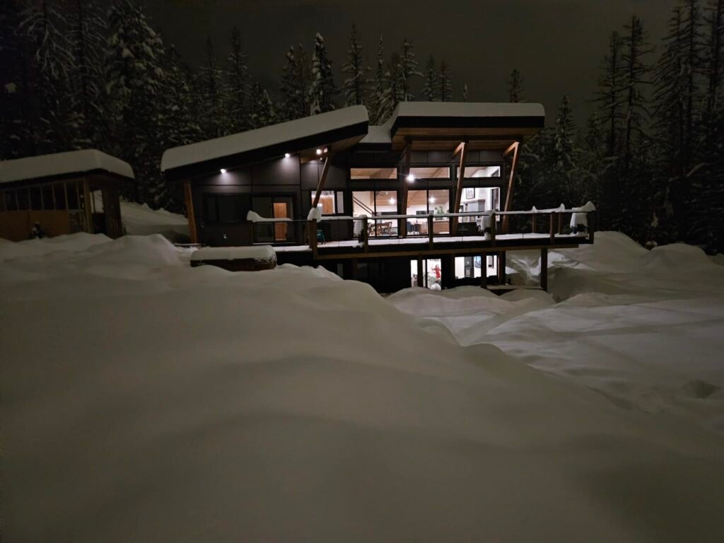 Exterior of a modern custom home built by DCB Construction glowing at night, surrounded by deep winter snow and forest in the East Kootenays.