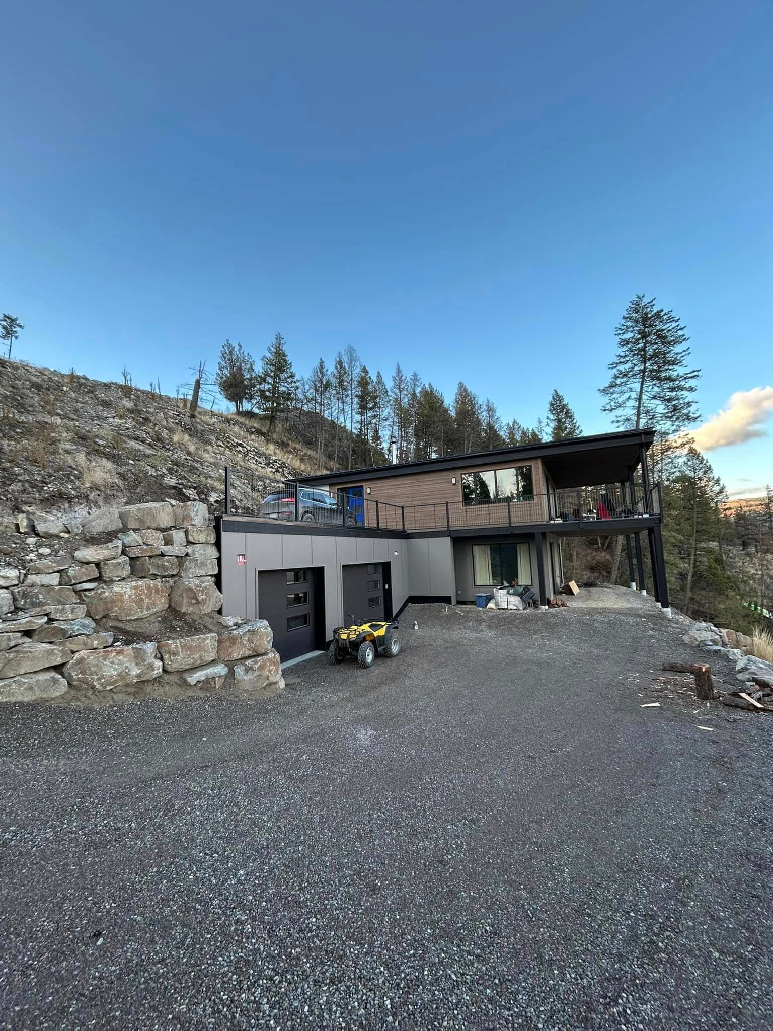 Exterior photograph of a custom modern carriage house built into a steep rocky hillside in the Kootenays featuring a lower level two car garage and an upper level living suite with a large covered deck.